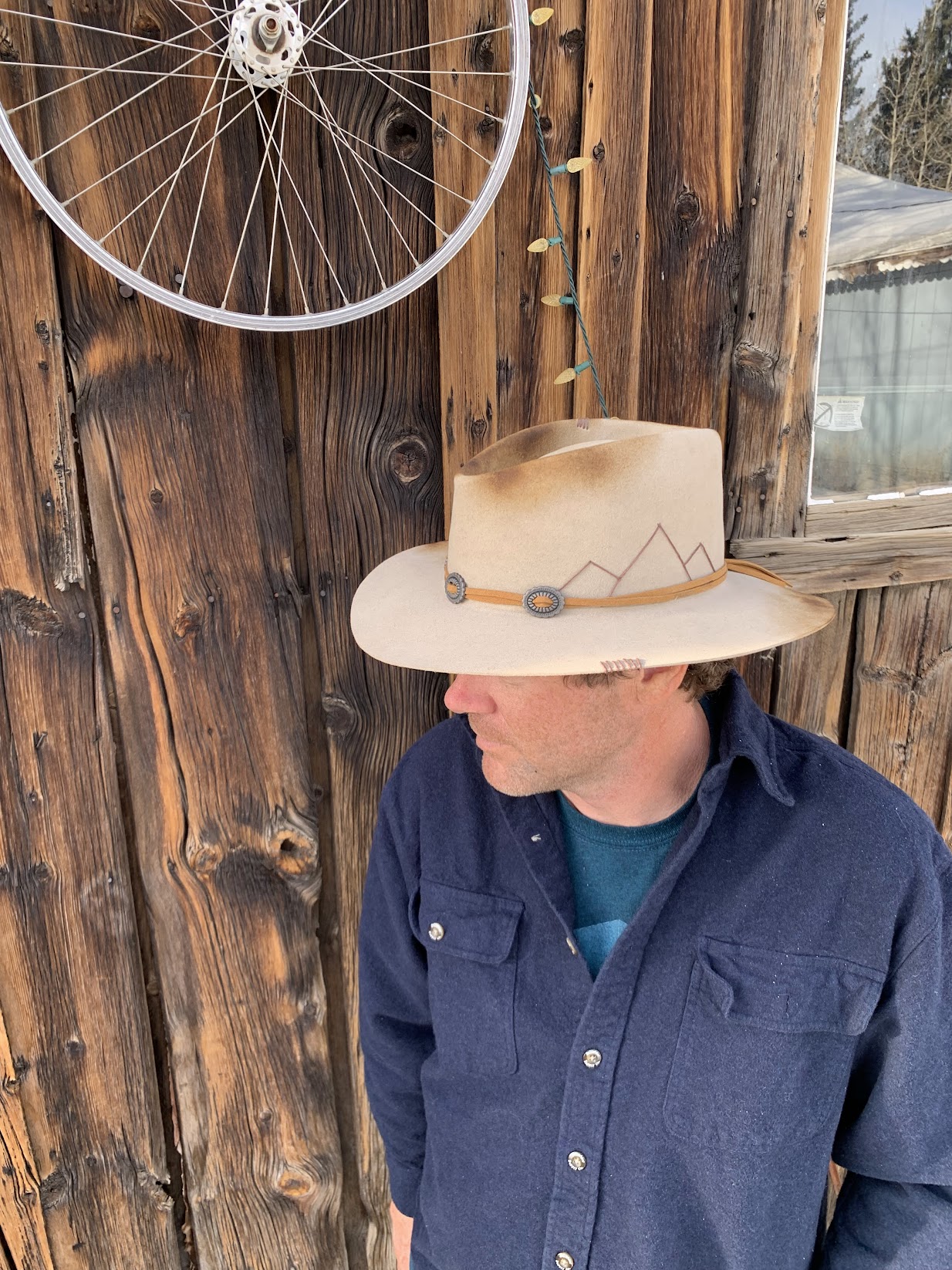 Adam Hevenor in a wide-brim hat, leaning against a weathered wood shed